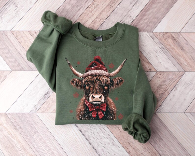 May include: Green sweatshirt with a brown and white highland cow wearing a red Santa hat and a red bow tie. The cow is surrounded by white snowflakes.