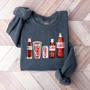 May include: A dark grey sweatshirt with a graphic of five different Coca-Cola products, including a glass bottle, a can and a plastic bottle. The text "Diet Coke" is visible on the graphic.