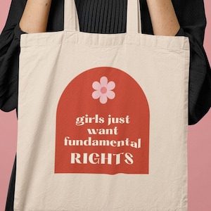 Può includere: Una borsa di tela bianca con un arco rosso e un fiore rosa. Il testo sulla borsa recita "girls just want fundamental RIGHTS".