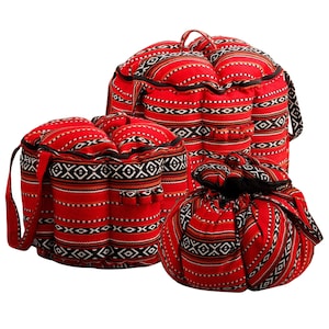 Puede incluir: Un conjunto de bolsas y cojines estampados en rojo y negro. Las bolsas tienen asas y cremalleras, mientras que los cojines son redondos e inflados. La tela presenta un diseño geométrico con acentos blancos, negros y marrones. Los artículos parecen estar hechos de una tela duradera.