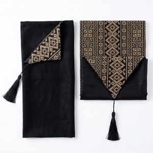 May include: Two black rectangular fabric runners featuring a gold-coloured geometric cross-stitch pattern along the edges and centre. Each runner is finished with a black tassel attached to a pointed corner, showcasing a solid black fabric base.