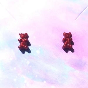 May include: A pair of red glitter gummy bear stud earrings. The earrings are small and detailed, with a shiny, textured surface. The earrings are set against a soft, pastel background.