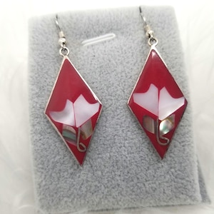 Peut inclure: Paire de boucles d'oreilles en forme de losange avec un fond rouge et un motif floral blanc. Les boucles d'oreilles ont des crochets argentés et des incrustations de coquillages irisés. Présentées sur une surface grise.