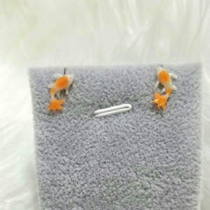 May include: Goldfish earrings. Each earring has a small, orange and white fish design. The earrings are presented on a grey, textured surface with a white clasp.