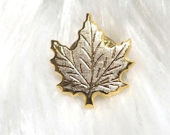 May include: A gold-colored maple leaf pin. The pin is a detailed representation of a maple leaf, with visible veins and a textured surface. The pin is likely made of metal and has a standard pin backing.