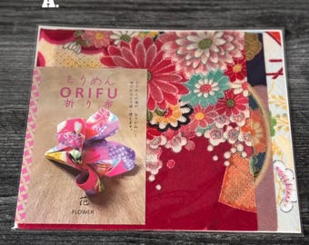 Puede incluir: Una hoja de tela cuadrada con un vibrante estampado floral en tonos rojos, rosas y blancos, superpuesta con una tarjeta rectangular etiquetada ORIFU con texto en japonés y una imagen de una flor de tela plegada etiquetada FLOWER.