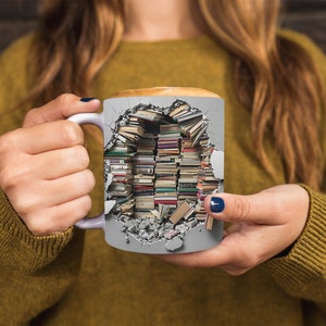 K&ouml;nnte beinhalten: Eine wei&szlig;e Keramiktasse mit einem Design einer zerbrochenen Wand, aus der B&uuml;cher herausbrechen. Die Tasse wird von den H&auml;nden einer Person gehalten.