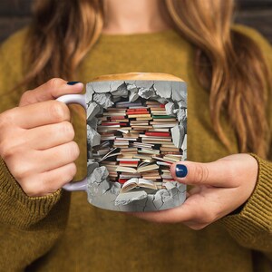 K&ouml;nnte beinhalten: Eine wei&szlig;e Keramiktasse mit einem Design aus einer zerbrochenen Backsteinmauer mit einem Stapel B&uuml;cher darin. Die Tasse wird von den H&auml;nden einer Person gehalten.