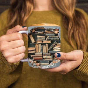 K&ouml;nnte beinhalten: Eine wei&szlig;e Keramiktasse mit einem Design eines B&uuml;cherregals, das mit alten B&uuml;chern gef&uuml;llt ist, die aus der Seite herausbrechen. Die Tasse wird von den H&auml;nden einer Person gehalten.