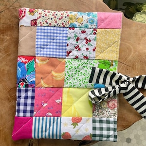 Puede incluir: Un salvamanteles acolchado de patchwork con una variedad de patrones de tela, incluyendo diseños florales, cuadros y frutas. El salvamanteles tiene un lazo a rayas blanco y negro adjunto en el lateral. El texto "Life is just a bowl of cherries" es visible.