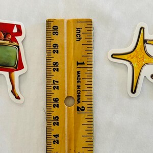 May include: Two die-cut stickers placed next to a wooden ruler. One sticker depicts a person in a red dress holding a vintage television set. The other sticker features three yellow, four-pointed star shapes with a glittery texture.