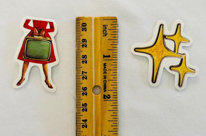 May include: Two die-cut stickers placed next to a wooden ruler. One sticker depicts a person in a red dress holding a vintage television set. The other sticker features three yellow, four-pointed star shapes with a glittery texture.