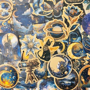 May include: A collection of celestial-themed stickers and cards. The designs feature moons, stars, planets, and mystical figures in shades of blue, gold, and cream. Some cards depict a castle, a cat, and a telescope.