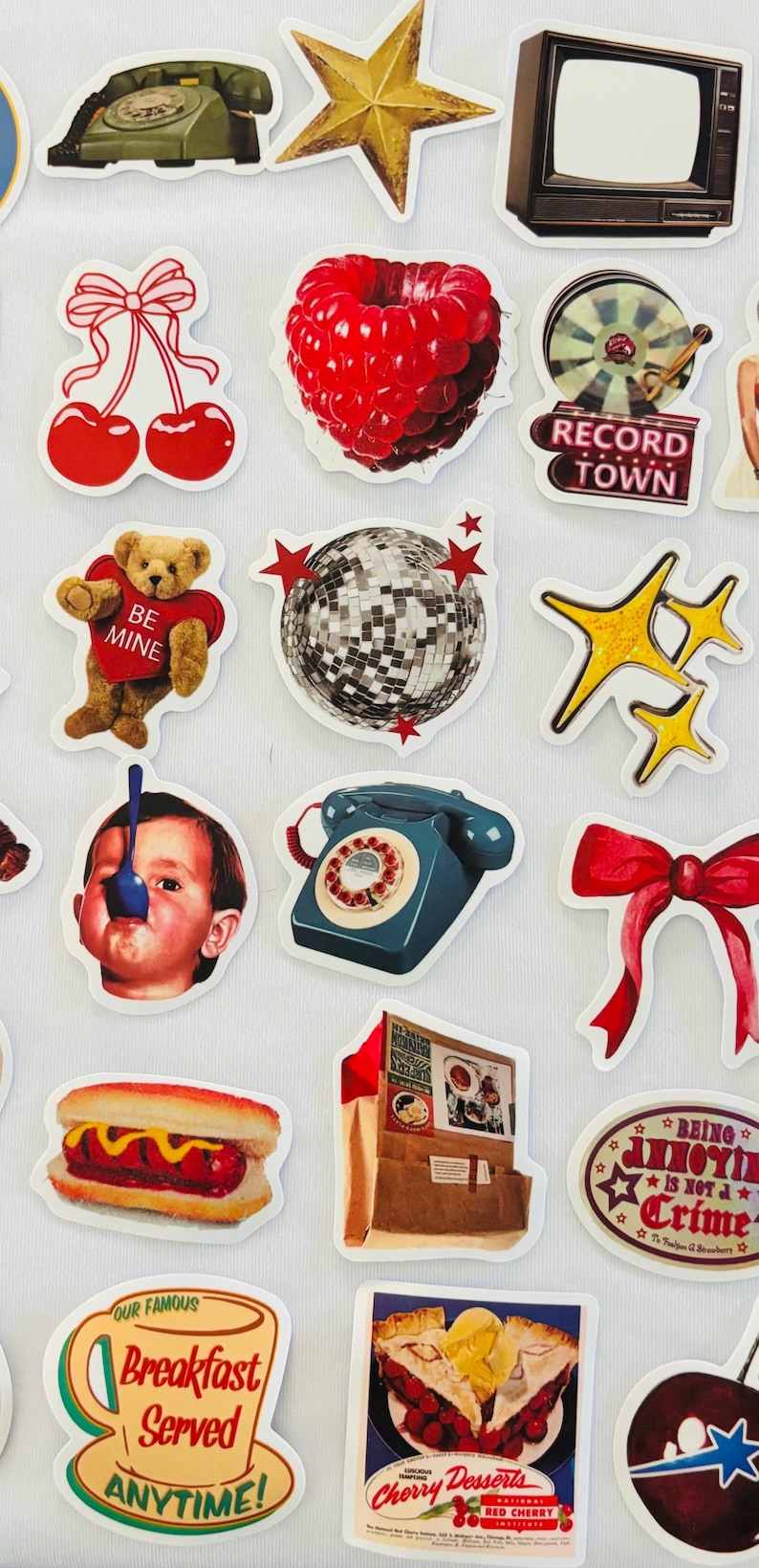 May include: A collection of assorted stickers featuring various illustrations including a green rotary phone, a gold star, a television, cherries, a raspberry, a record player, a teddy bear, a disco ball, a child with a spoon, and food items.