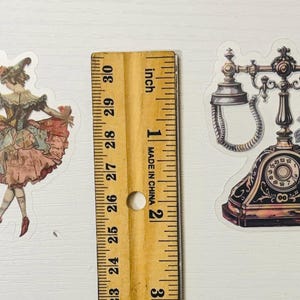 Pu&ograve; includere: Due adesivi in stile vintage: una ballerina con un vestito rosa e blu e un telefono antico nero e oro con rose rosa. Un righello di legno con misure in pollici &egrave; tra gli adesivi.