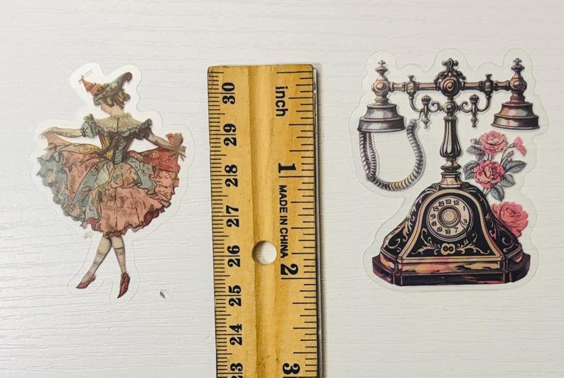 Pu&ograve; includere: Due adesivi in stile vintage: una ballerina con un vestito rosa e blu e un telefono antico nero e oro con rose rosa. Un righello di legno con misure in pollici &egrave; tra gli adesivi.