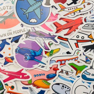 May include: A collection of colorful airplane stickers in various designs. The stickers feature different types of aircraft, including jets and propeller planes, in shades of blue, red, pink, and green. Some stickers have text like "PILOTS" and "LET DOWN ON PEOPLE".