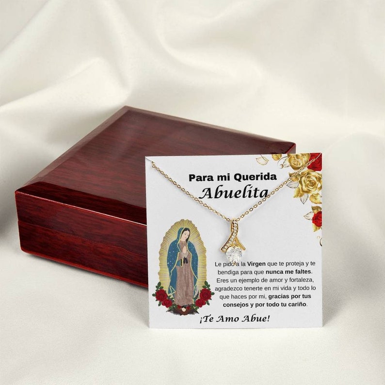 May include: A gold necklace with a pendant featuring the Virgin Mary and a small, clear crystal. The necklace is displayed in a wooden box with a white card that reads "Para mi Querida Abuelita" and a message about the Virgin Mary.