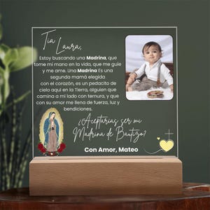 May include: A personalized baptism gift featuring a photo of a baby, text in Spanish, and an image of the Virgin of Guadalupe. The clear acrylic plaque sits on a wooden base. The text includes the phrase "Tia Laura".