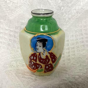 May include: A small, vintage ceramic vase with a green top and a cream-coloured body. The vase features a painted portrait of a person in a blue circle, wearing a red and white patterned garment. The vase is approximately 10 cm tall.