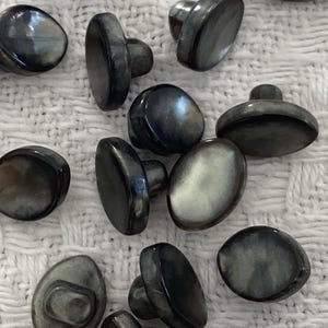 May include: A collection of 18 oval-shaped, black and grey shell buttons with a raised centre post. The buttons have a smooth, glossy finish.