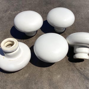 May include: Five white ceramic mushroom-shaped cabinet knobs. The knobs have a glossy finish and a rounded top. One knob has a visible screw hole. Suitable for furniture restoration or DIY projects.