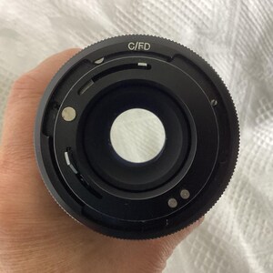 May include: Close-up of a black camera lens with the text "C/FD" on the outer ring. The lens has a circular opening and several small metal pins. The lens is held against a white, textured background.