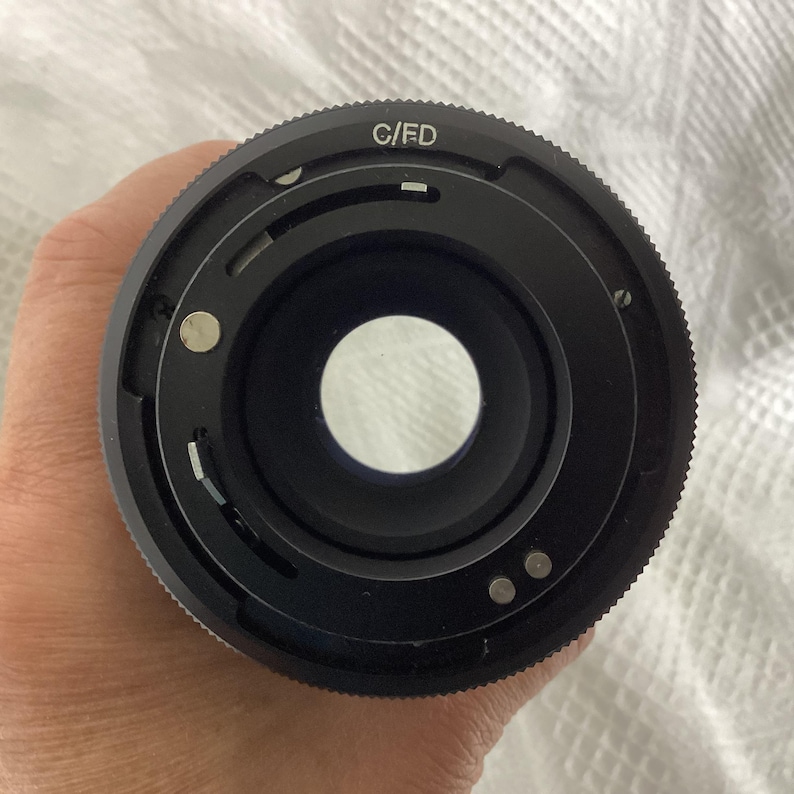 May include: Close-up of a black camera lens with the text "C/FD" on the outer ring. The lens has a circular opening and several small metal pins. The lens is held against a white, textured background.