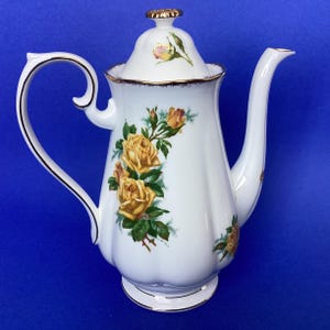 May include: White porcelain coffee pot with a lid, handle, and spout. The pot is decorated with yellow roses and green leaves. Gold trim accents the edges of the pot, lid, and handle. The pot sits on a small, round base.
