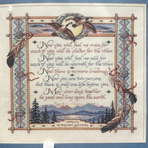 May include: A cross-stitch pattern featuring a blue and green landscape scene with a quote about marriage. The quote reads: "Now you will feel no rain for each of you will be shelter for the other. Now you will feel no cold for each of you will be warmth for the other. Now there is no more loneliness. Now you are two persons but there is only one life before you. May your days together be good and long upon the earth." The pattern is titled "Apache Marriage Blessing."
