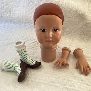 May include: A porcelain doll head with brown hair, freckled cheeks, and closed eyes. The doll head is separate from the body and is accompanied by two porcelain arms and two porcelain legs with green and white striped socks.