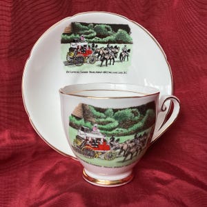 May include: A white teacup and saucer set with gold edging. The set has a colourful illustration of a horse-drawn carriage on a road, with text reading "BX Express, Cariboo Road, About 1965, Williams Lake, BC."