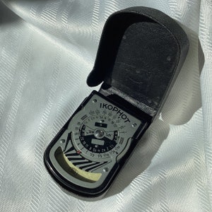 Peut inclure: Un posemètre noir et argent avec un étui en cuir. Le posemètre a un cadran avec les mots "IKOPHOT", "DIN" et "Sch" dessus. Le cadran comporte des chiffres de 1 à 60 et de 1/2 à 1/25.