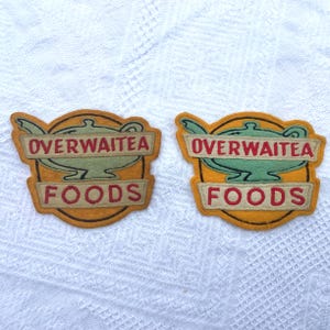 May include: Two felt patches with the text "OVERWAITEA FOODS". The patches are yellow with a green teapot design and red lettering. The patches are on a white, textured surface.
