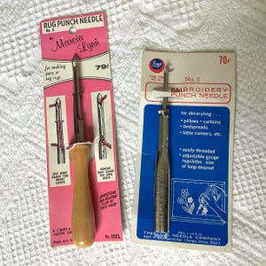 May include: Two vintage rug punch needles on their original packaging. One is pink with the text "RUG PUNCH NEEDLE" and "Marcia Lynn." The other is blue with "EMBROIDERY PUNCH NEEDLE" and "Boye." Both are for making decorative items.