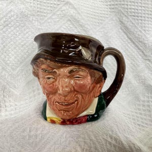 May include: A ceramic jug in the shape of a smiling man's face, with a brown hat and a green jacket, and a red bow tie. The handle is brown, and the jug has a glossy finish.