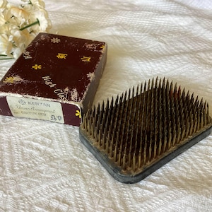 May include: A rectangular Kenzan flower arranging tool with brass pins set in a dark gray base. A brown box with gold lettering and floral designs is next to the Kenzan. The box reads "High Class" and "Flower Arrangement".