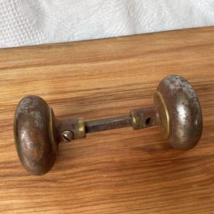 May include: A vintage door knob set featuring two weathered, tarnished metal knobs connected by a central square metal spindle. The knobs have a dark, oxidised patina with visible surface wear, and each is attached to a brass-toned circular base plate.