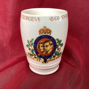 May include: White ceramic mug with a royal crest featuring King George VI and Queen Elizabeth. The crest is surrounded by oak leaves and acorns. The text "HONI SOIT QUI MAL Y PENSE" is written around the crest. The text "DIEU ET MON DROIT" is written below the crest.
