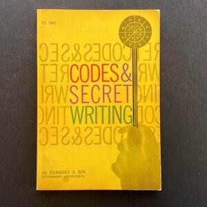 May include: A yellow paperback book titled Codes & Secret Writing by Herbert S. Zim, marked TX 164. The cover features a circular cipher wheel graphic, a vertical line, and a silhouette of a hand, with the title text in red, purple, and green.