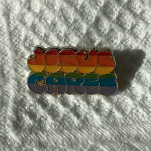 May include: A rainbow-coloured enamel pin with the text "JESUS CARES". The pin features a silver border and is set against a white, textured backdrop. The letters are in a rounded, playful font.