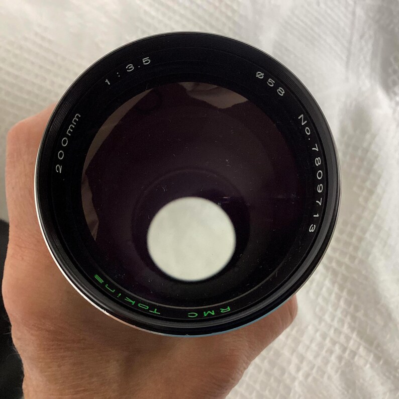 May include: A black Tokina RMC 200mm camera lens with a 58mm diameter. The lens has the markings "1:3.5" and "No. 7809713" printed on the black metal casing. The lens is held in a hand.