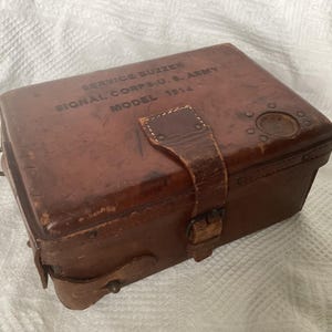May include: A vintage brown leather box with a buckle closure and a leather strap. The text "SERVICE BUZZER SIGNAL CORPS U.S. ARMY MODEL 1914" is printed on the top. The box has a metal ring on the side.