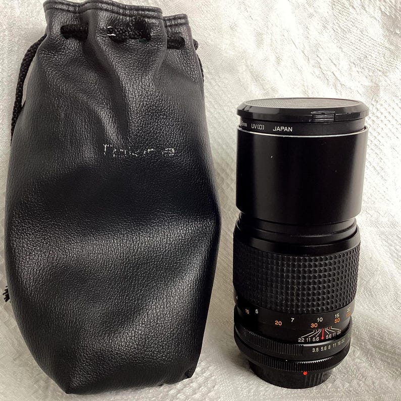 May include: Black camera lens with a textured grip and measurement markings, next to a black leather drawstring pouch. The lens has a black cap with the text "UV(01) JAPAN". The pouch has the text "Tokina" printed on it.