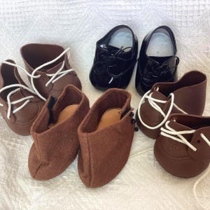 May include: A selection of doll shoes in different styles and colours. The collection includes brown leather oxfords with white laces, black patent leather dress shoes, and brown felt booties. The shoes are arranged on a white textured surface.