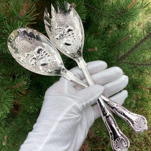 May include: A pair of silver-plated salad servers featuring ornate embossed fruit and leaf motifs on the bowls. The handles are decorated with a shell-like pattern at the base. The polished metal surfaces reflect light, highlighting the intricate details.