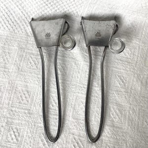 May include: Two vintage silver-toned metal ice tongs. Each tong features a small, rectangular bucket-shaped head with a decorative emblem and a clear glass ring. The long, curved handles are connected to the head.