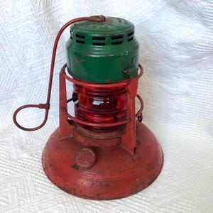 Peut inclure: Une lanterne en métal vintage dotée d'une base circulaire rouge vif et d'un cadre en fil de fer rouge assorti. La section centrale contient un globe en verre rouge nervuré, surmonté d'un capuchon ventilé vert. Une poignée en métal rouge est fixée.