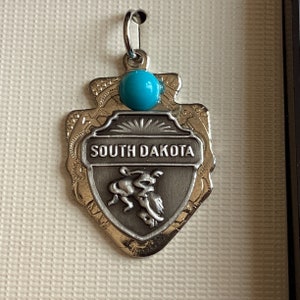 May include: A silver charm in the shape of a shield with the words "South Dakota" and a stylized image of a buffalo. The charm has a turquoise blue bead at the top.