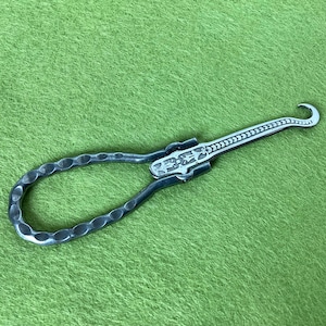 May include: A silver-toned metal button hook featuring a hammered, textured handle loop and a slender, engraved shaft that terminates in a curved hook. The tool displays intricate decorative patterns along the central hinge and the metal shank.